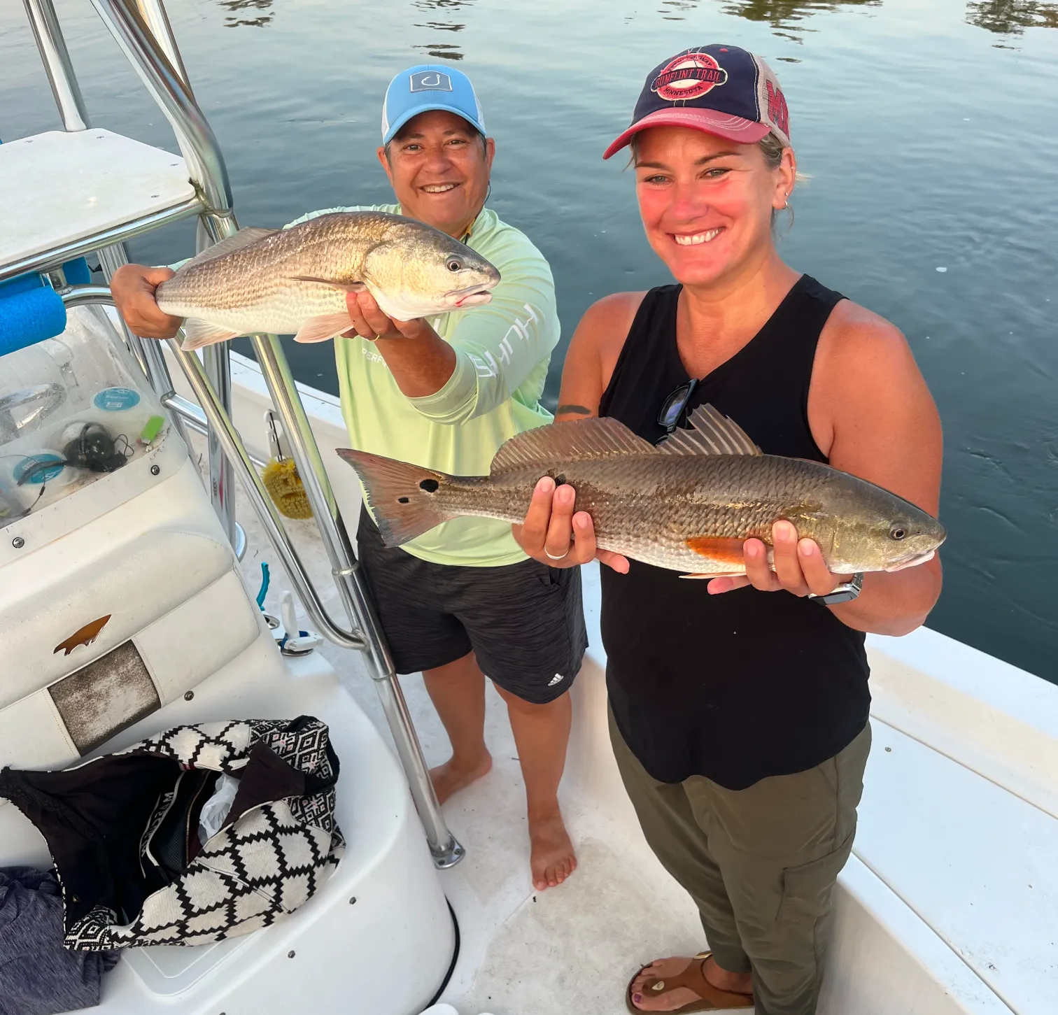 Two smiling people on a boat each holding a freshly caught fish over the water.