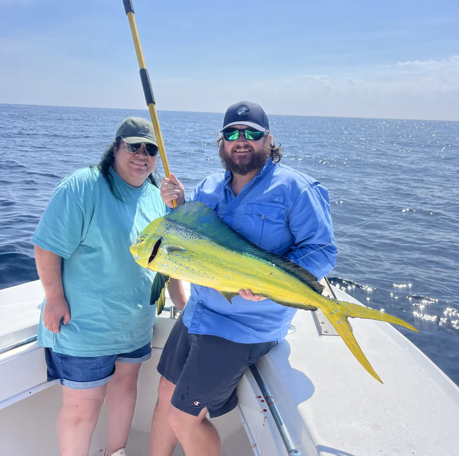 Two people on a boat hold a large, bright yellow fish with the ocean and blue sky in the background.