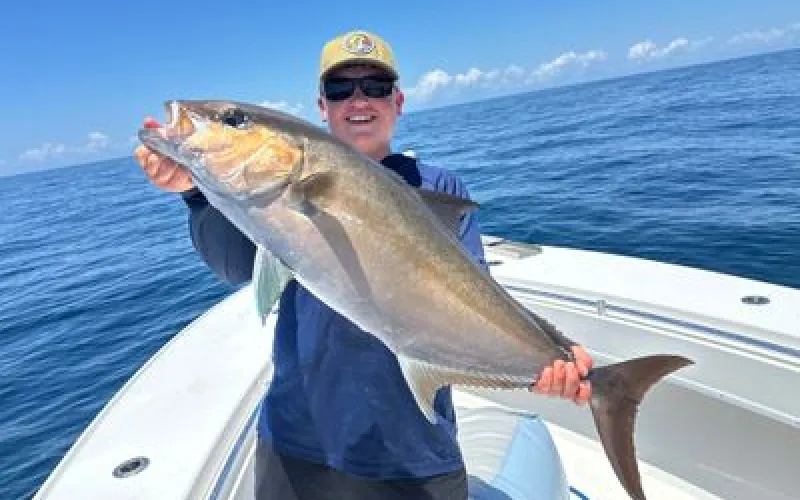 Person smiling and holding a large fish on a boat with the ocean and blue sky in the background.