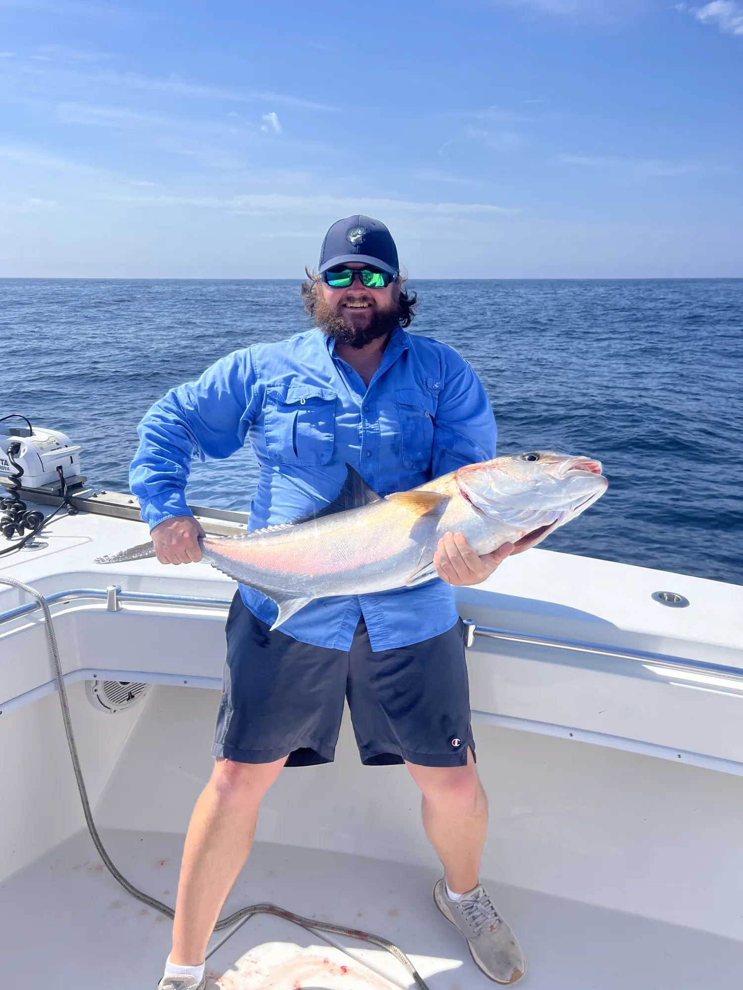 nearshore-w Man in blue shirt and hat holding a large fish on a boat with the ocean and blue sky in the background.