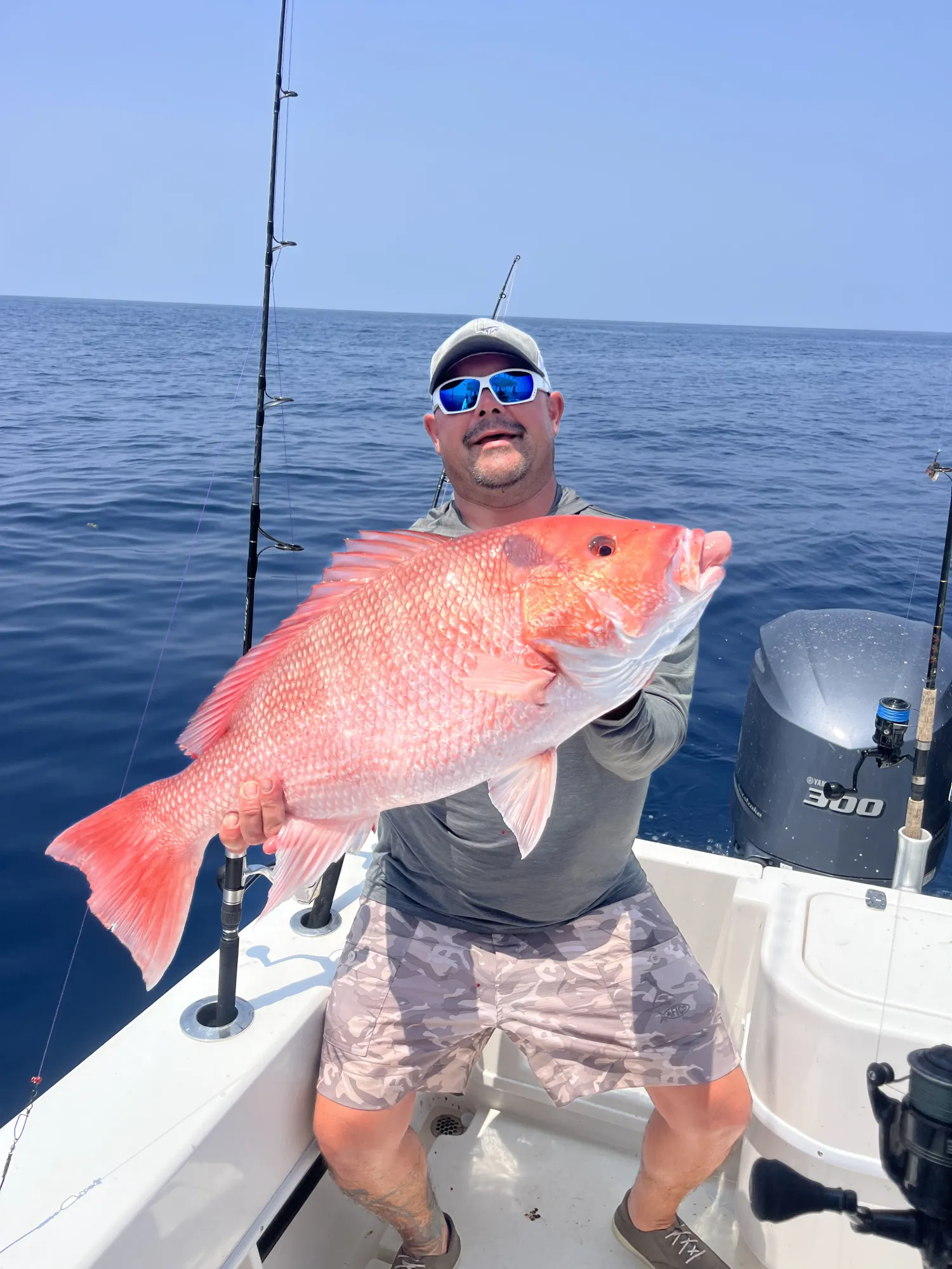 Man on a boat holding a large red snapper fish with ocean and fishing poles in the background.