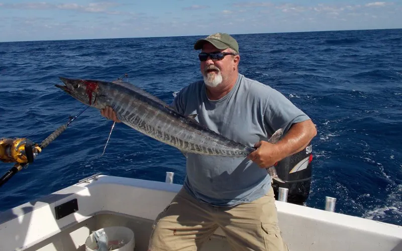 Man in sunglasses and cap holding a large fish on a boat in the ocean under a partly cloudy sky.