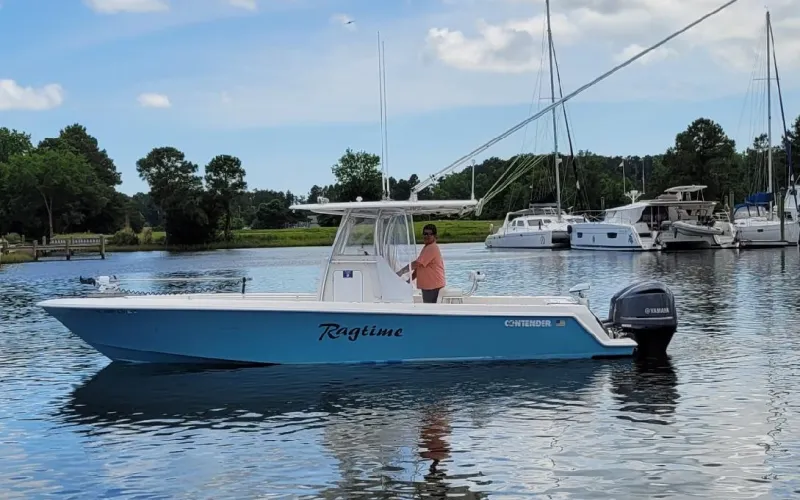 Person steering a blue and white boat named "Ragtime" on calm water with other boats and trees in the background.