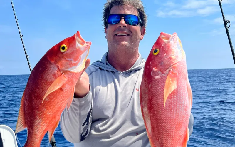 Smiling person on a boat holding two large red fish with the ocean and sky in the background.