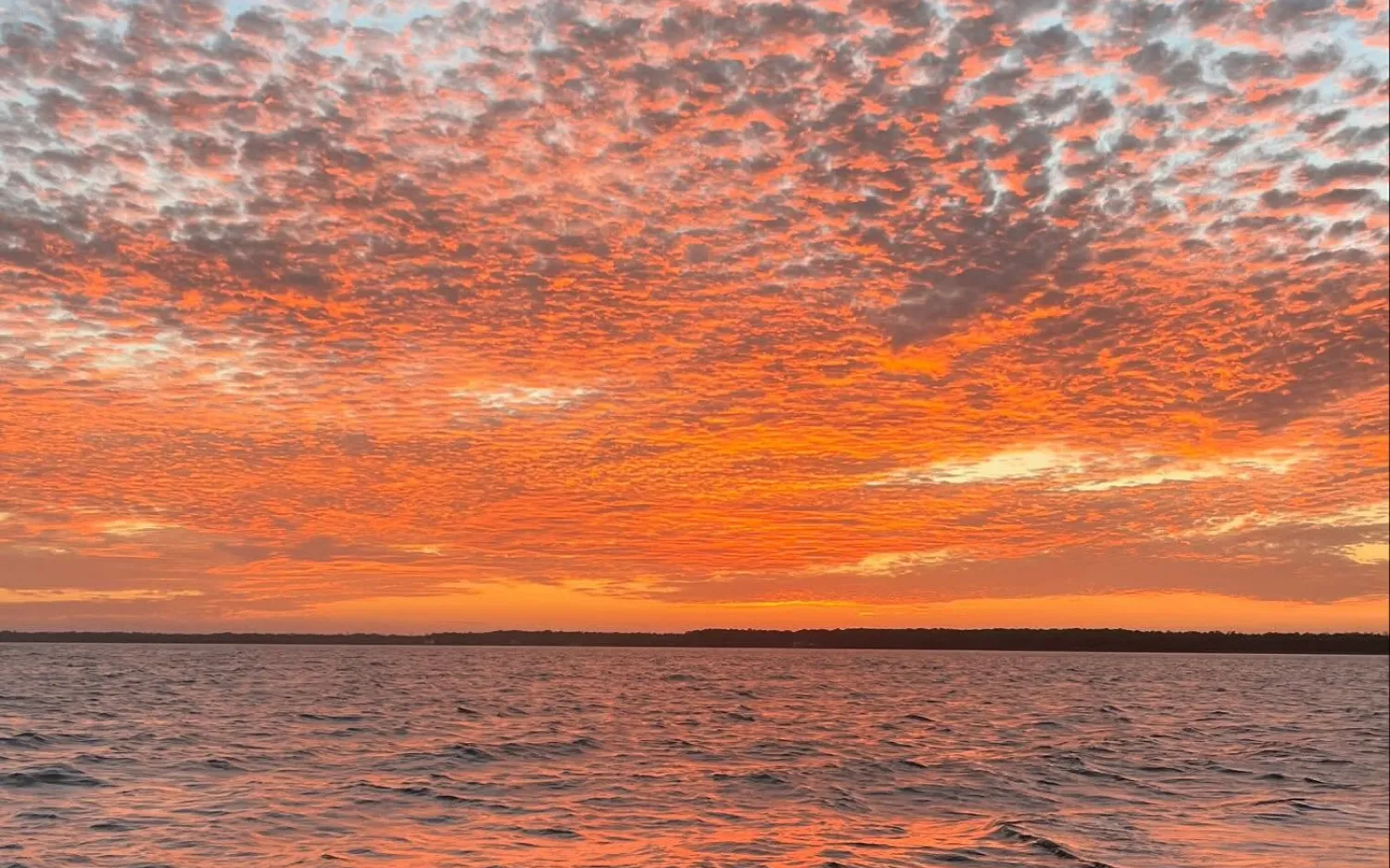 Vivid orange clouds fill the sky over a calm body of water at sunset, with a dark horizon line.
